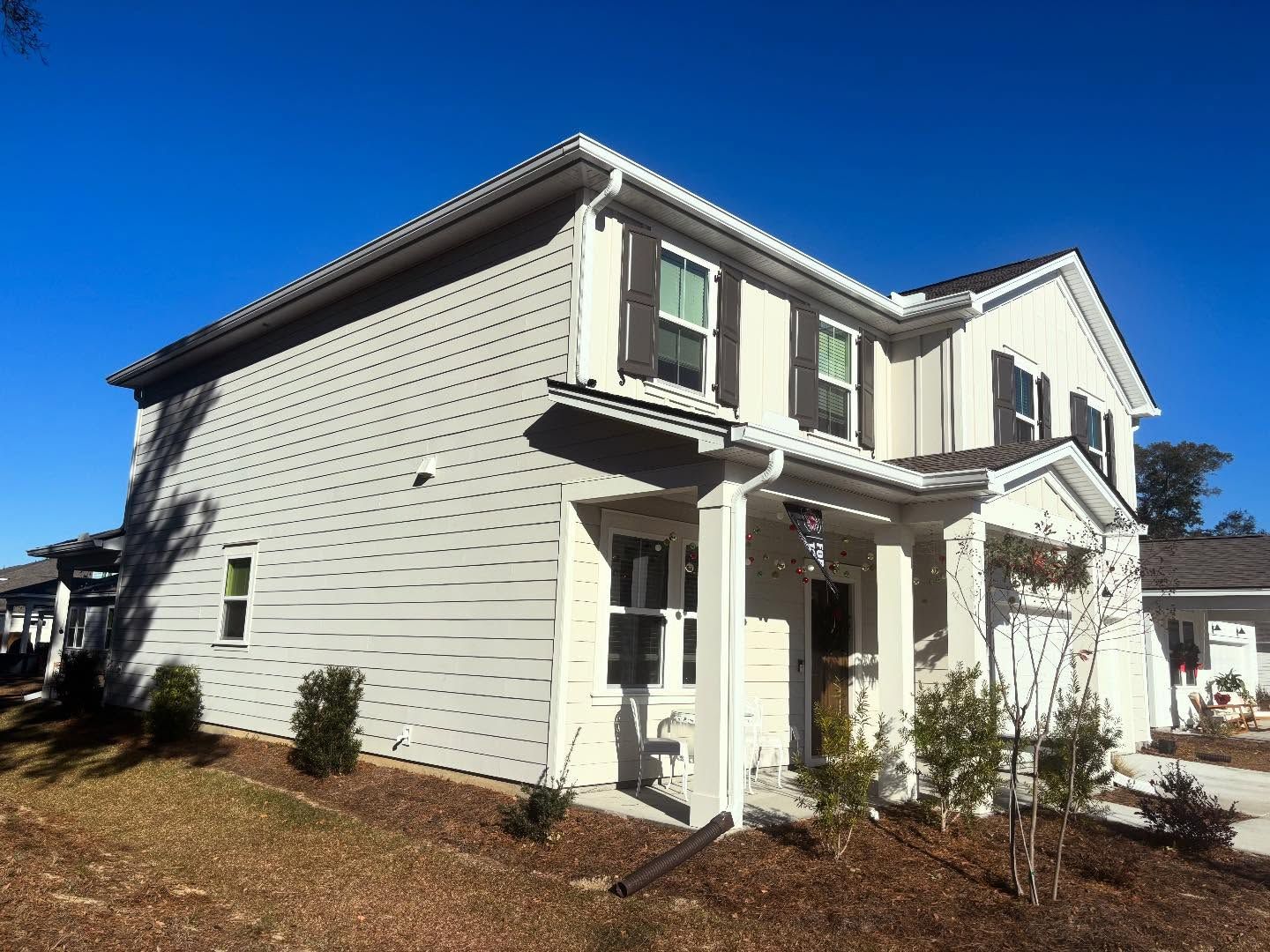 A two-story, light-colored suburban house with dark shutters, a covered porch, and landscaping under a clear blue sky.