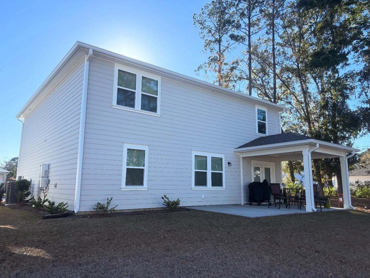 A two-story light gray house with a covered back patio, multiple windows, and a yard under a clear blue sky.
