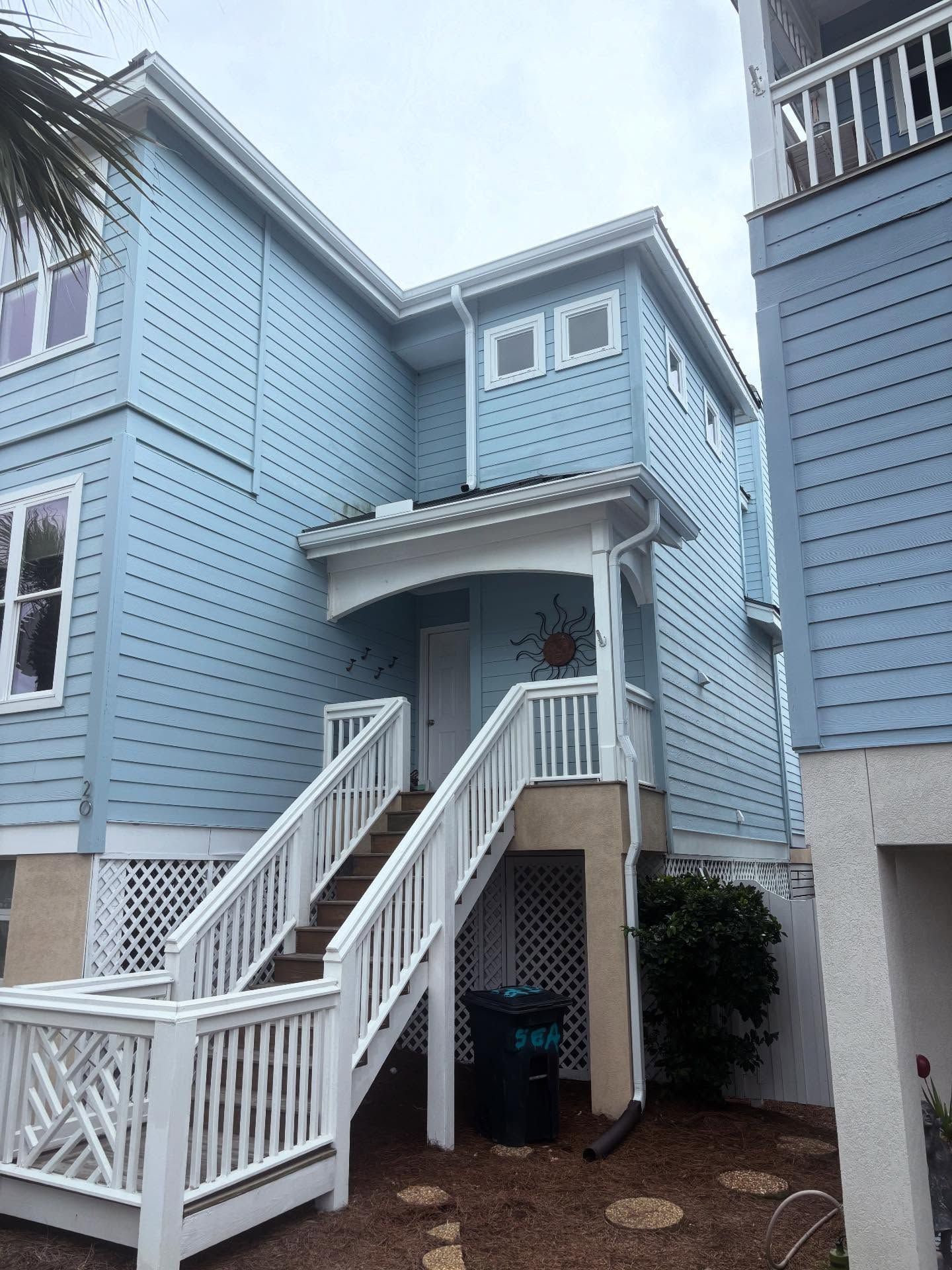 A light blue, multi-story beach house with elevated wooden stairs, a white railing, and a black trash bin underneath.