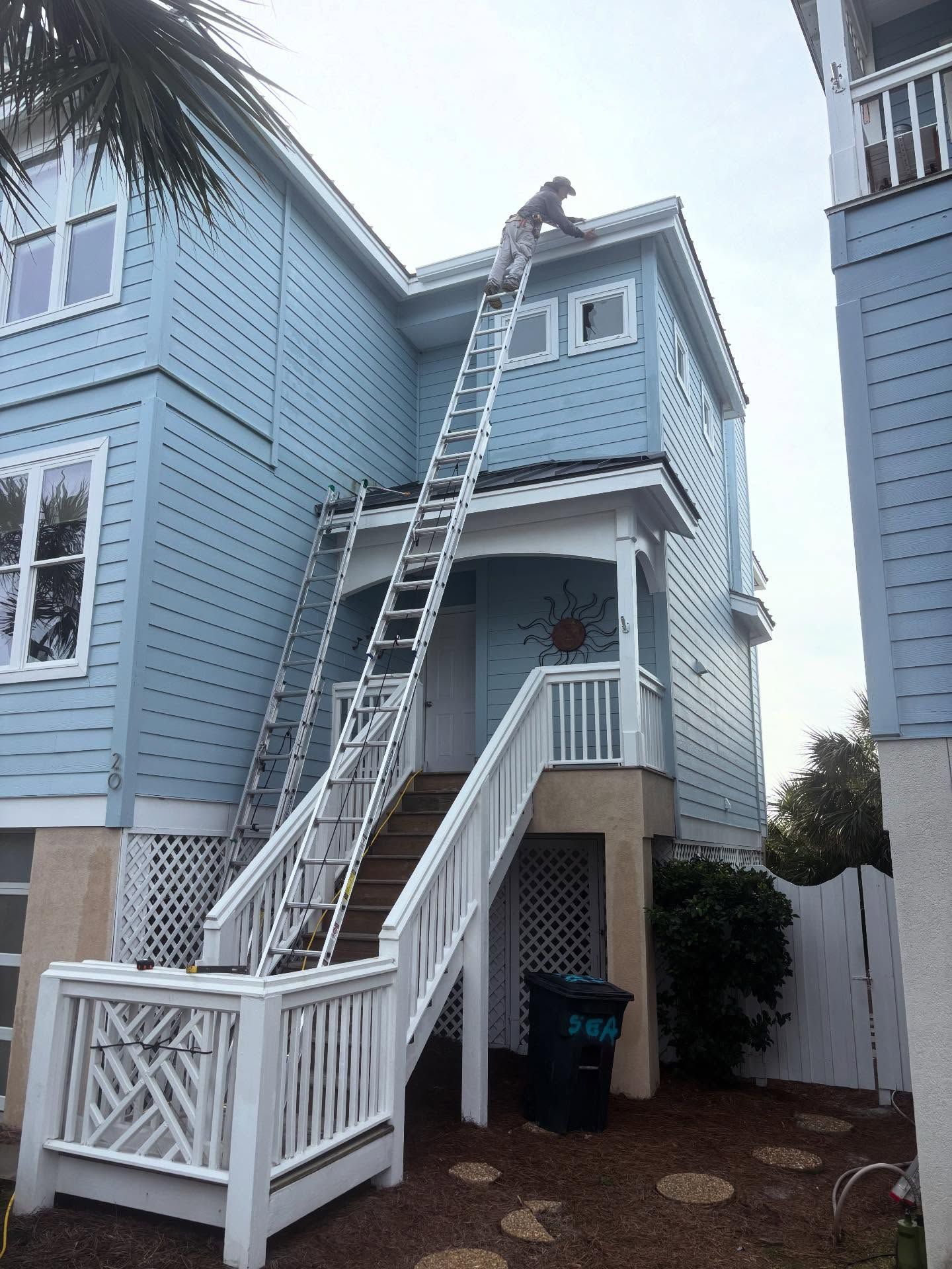 A worker on a tall extension ladder cleaning the gutters of a two-story light blue house with a white staircase.