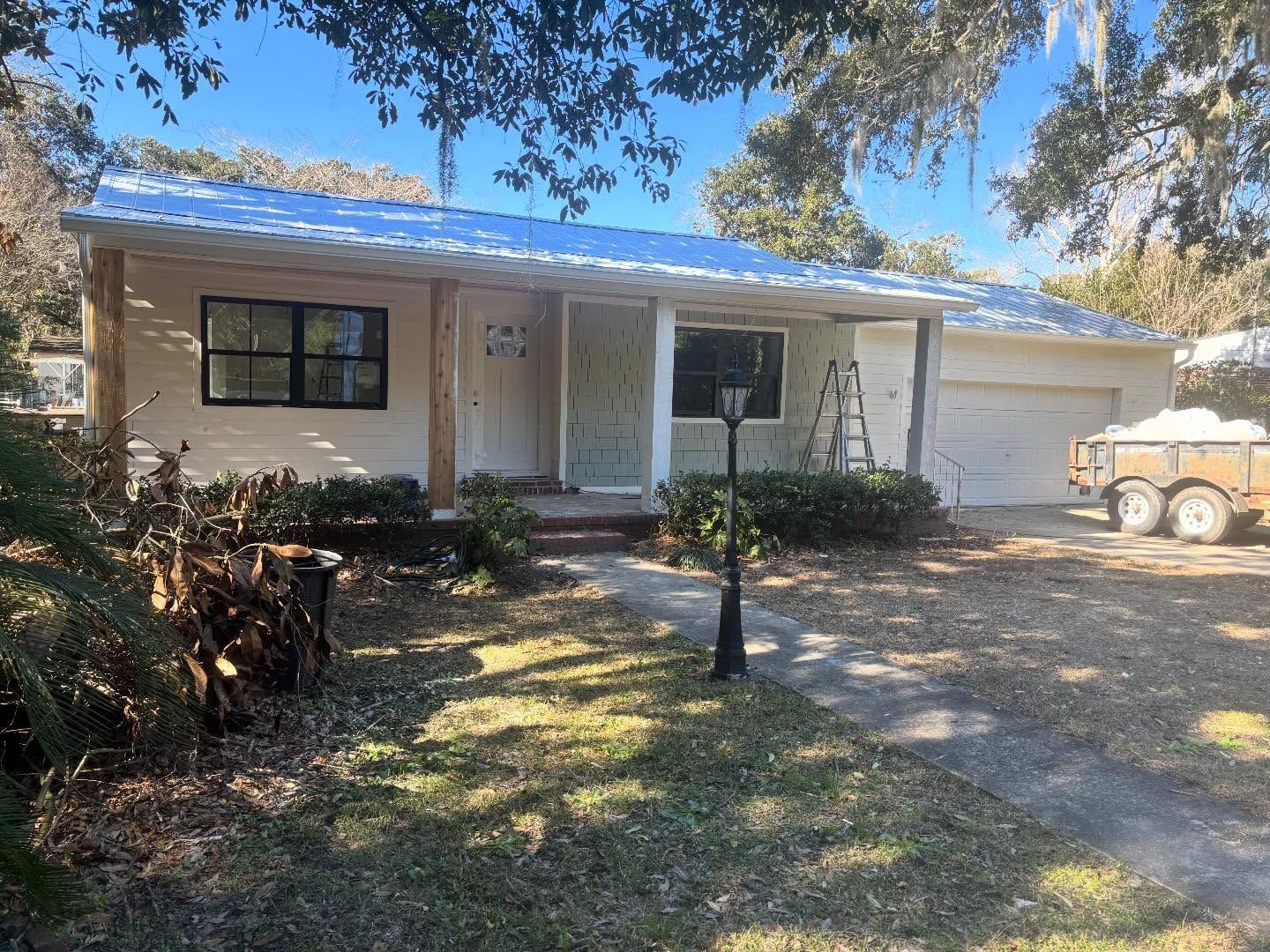 Single-story house with a white exterior, covered porch, and attached garage under a metal roof on a sunny, leafy lot.