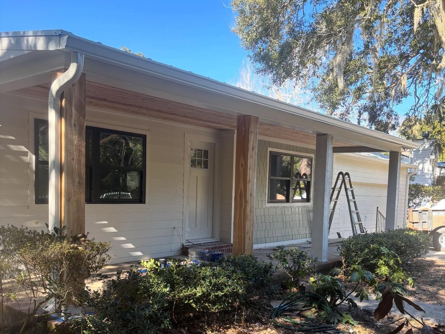 A beige, single-story house with a front porch, dark-framed windows, and unpainted wooden support posts.