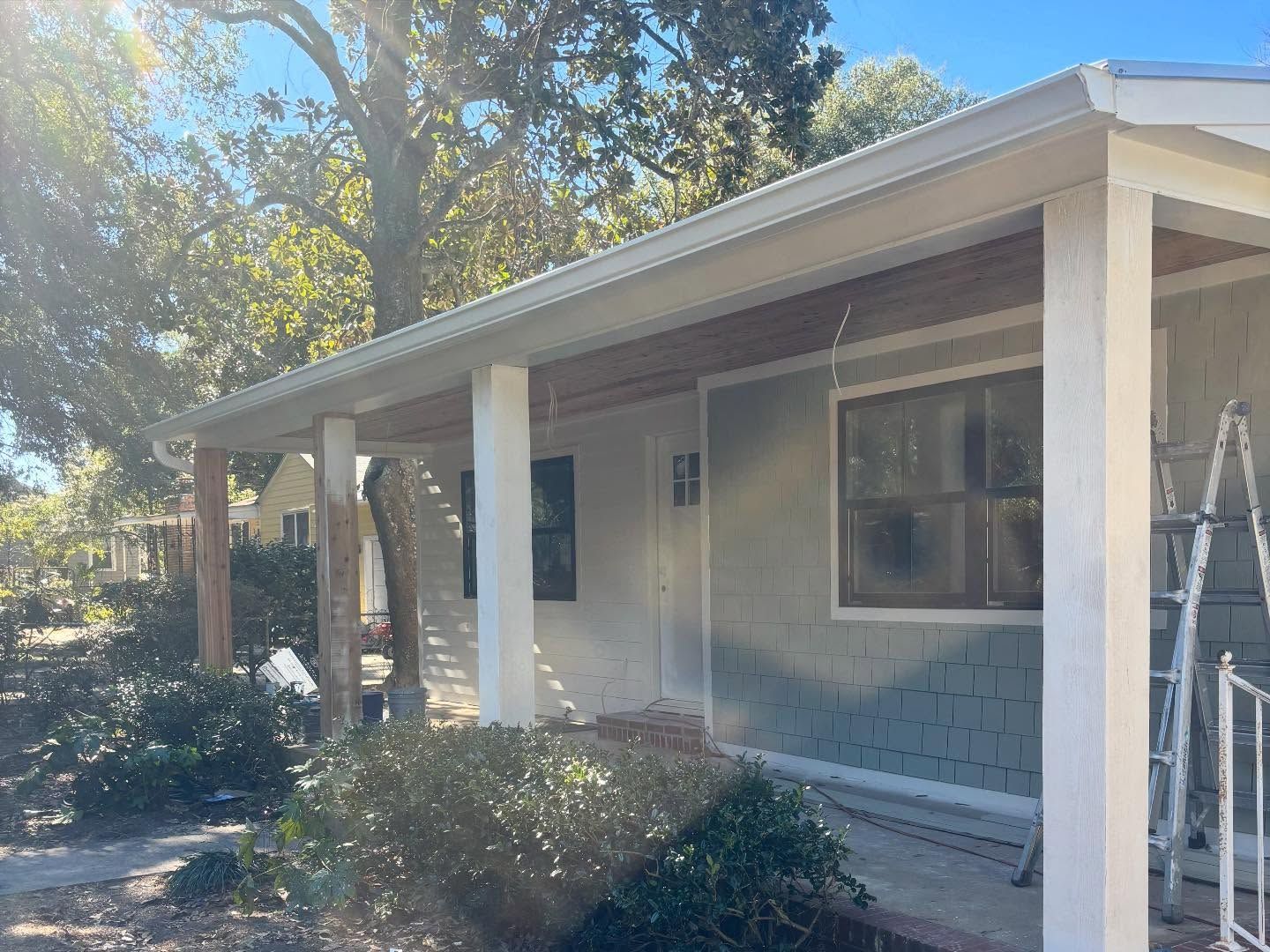 A light gray, one-story house with a white-pillared porch under a bright blue sky, with a ladder leaning against the wall.