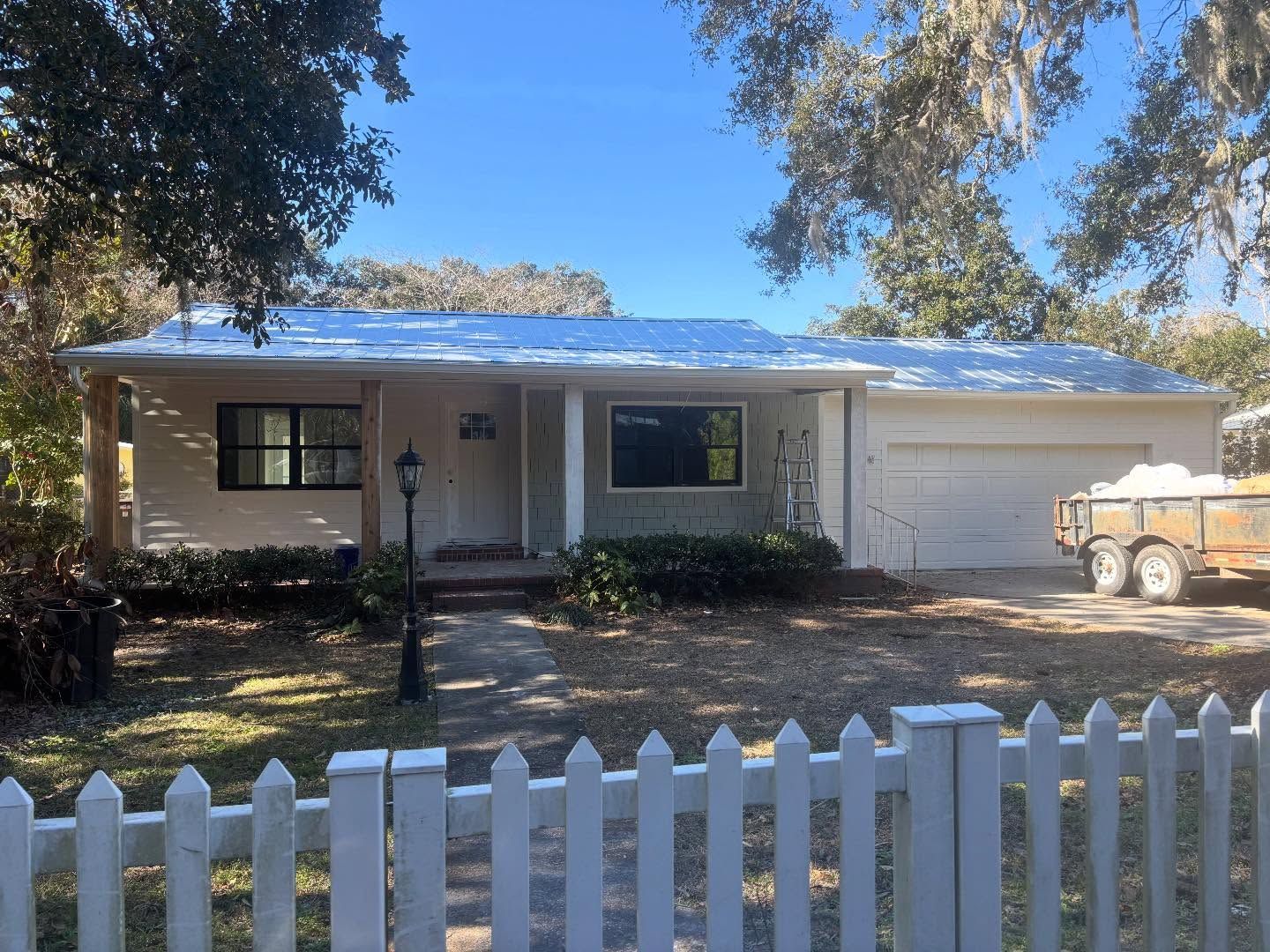 A single-story, light-colored house with a dark roof and garage, viewed behind a white picket fence under a clear blue sky.