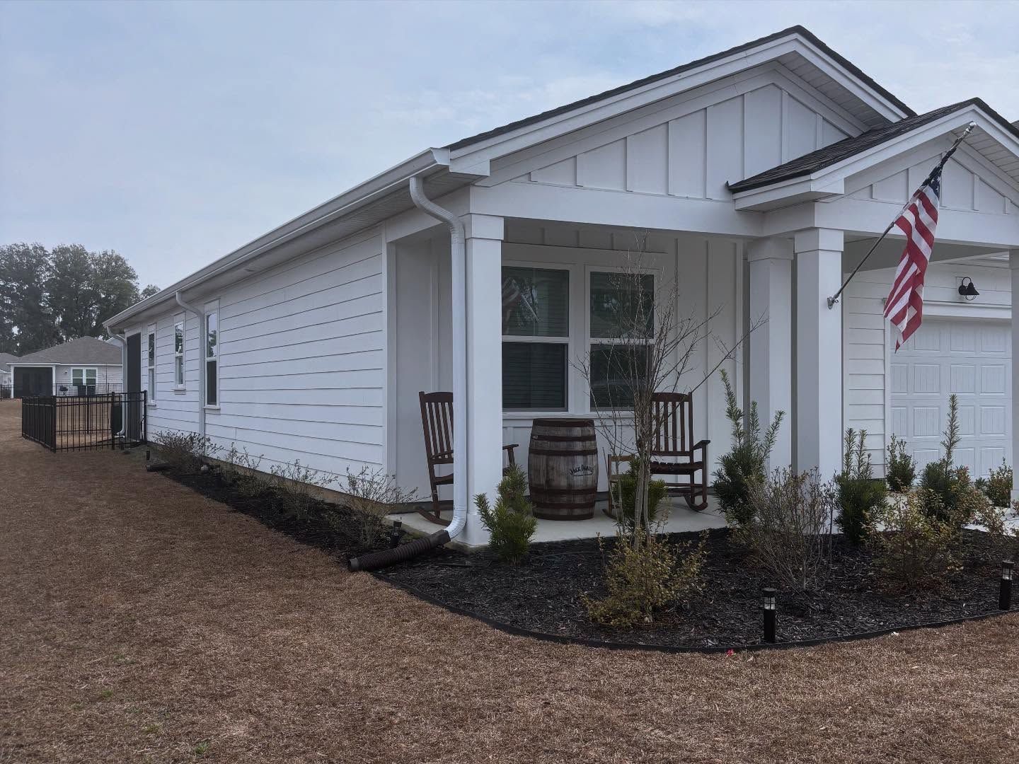 A white, single-story suburban house with a covered front porch featuring two chairs, a barrel, and an American flag.
