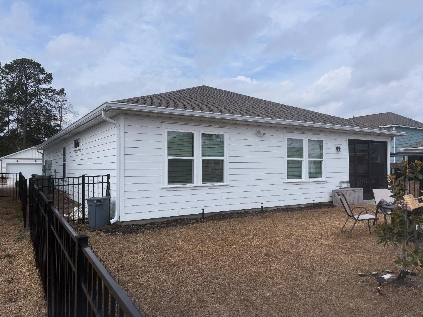 A white, single-story house with a grey roof and fenced backyard under a cloudy sky.