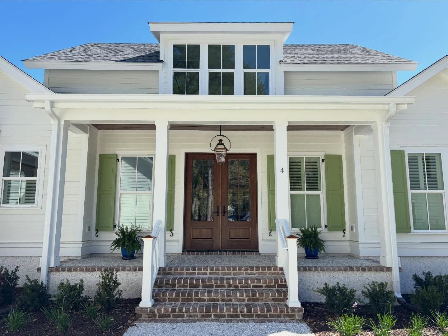 A white house exterior featuring a brick front porch, symmetrical green shutters, a brown double door, and a dormer window.