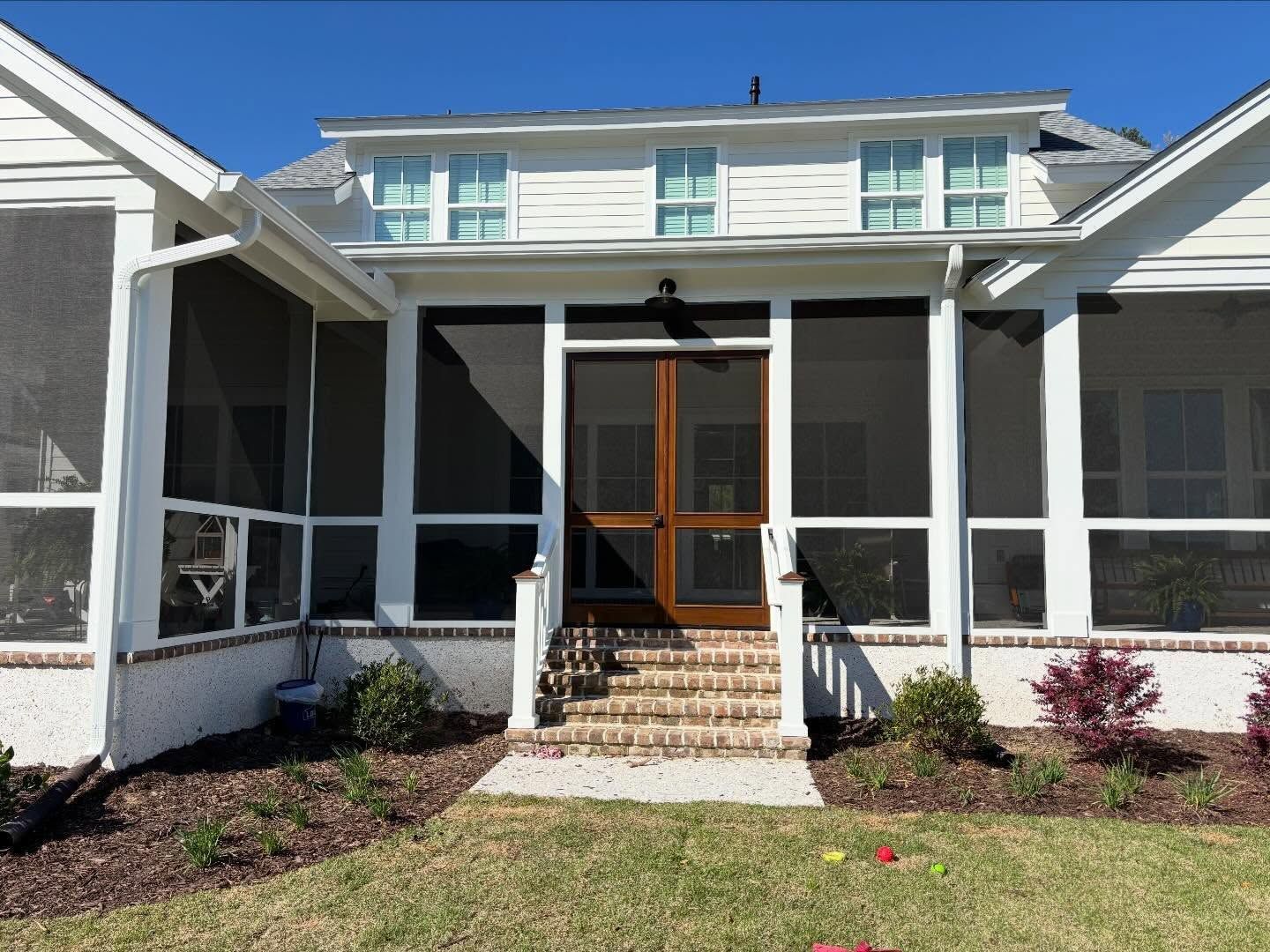 A white house with a screened-in back porch, wooden screen doors, brick stairs, and landscaped garden beds.
