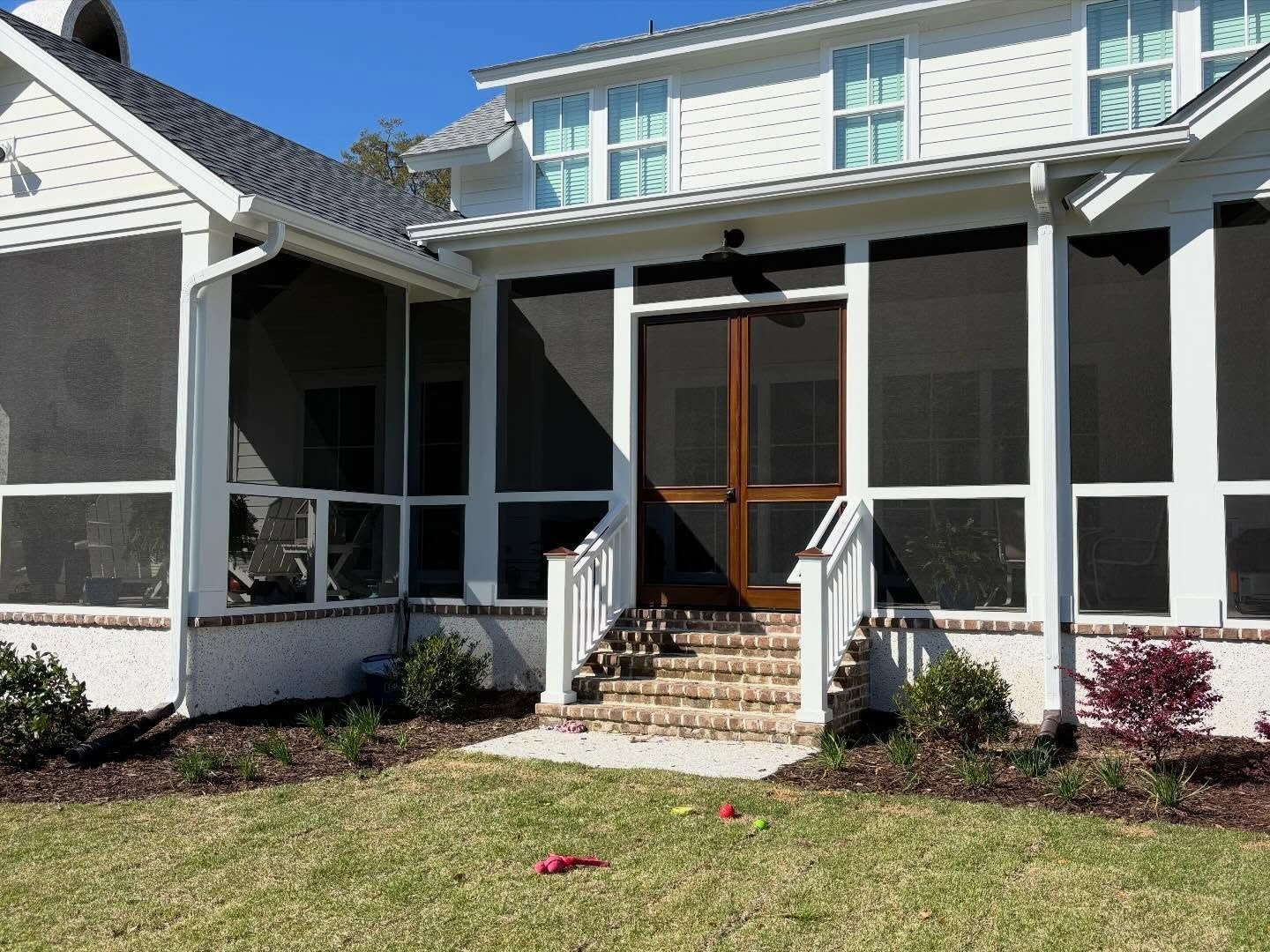 A white-sided home with a screened-in porch, brick steps leading to a wooden double door, and a landscaped lawn.