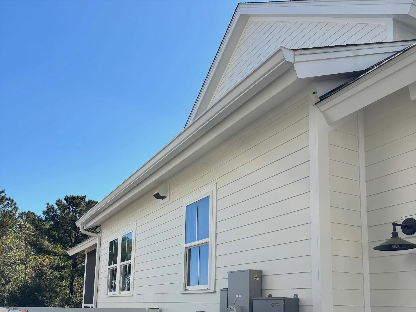 A light-colored house exterior with white siding, a metal roof, two windows, a light fixture, and a clear blue sky.