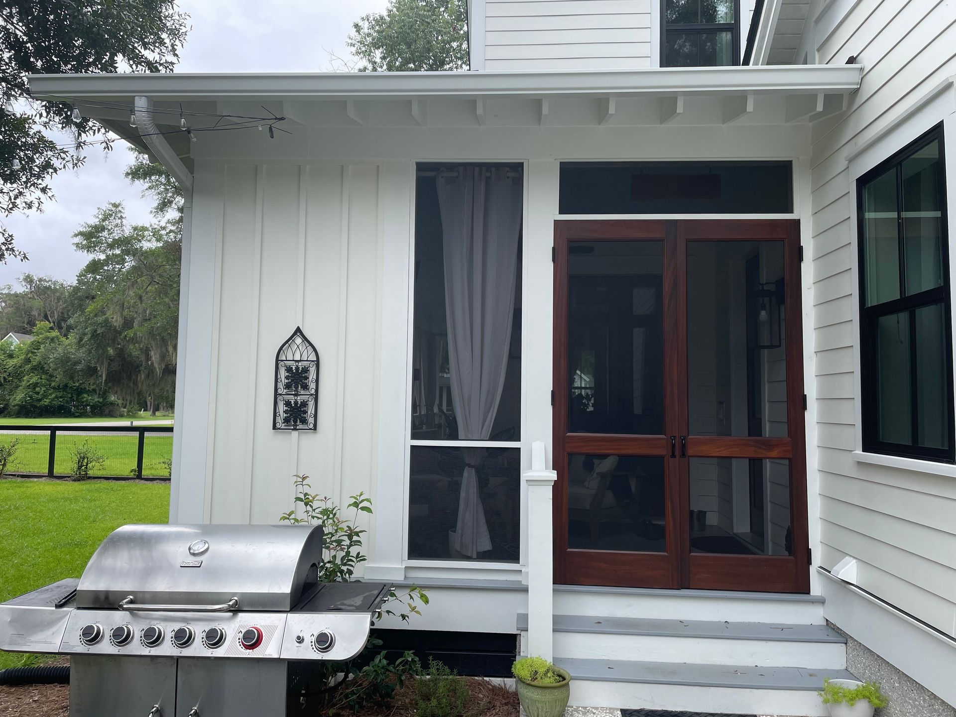 A stainless steel grill sits on a patio next to the wooden screen doors and white siding of a house.