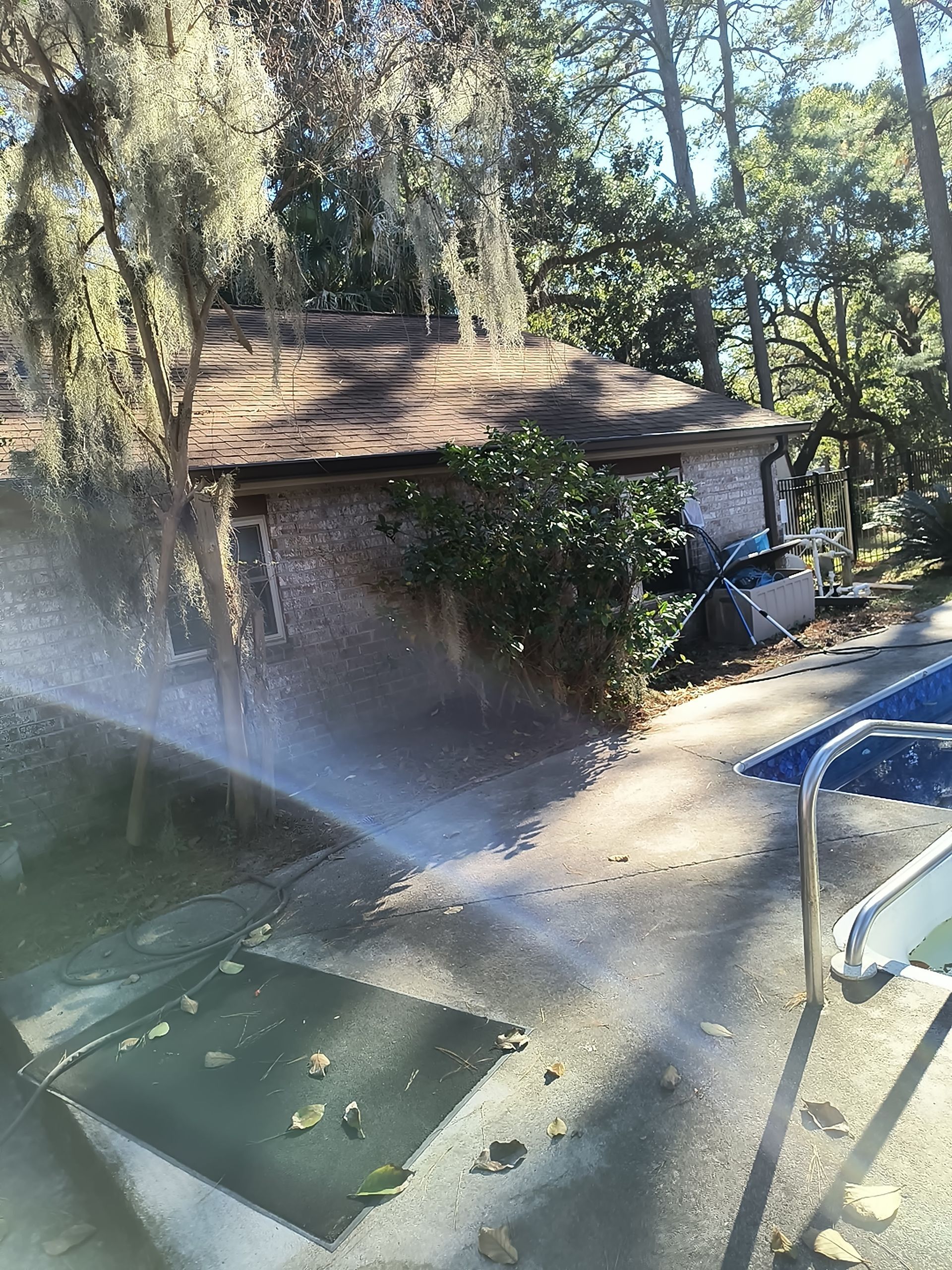 A high-angle view shows a sprinkler spraying water across a concrete patio next to a swimming pool and a brick house.