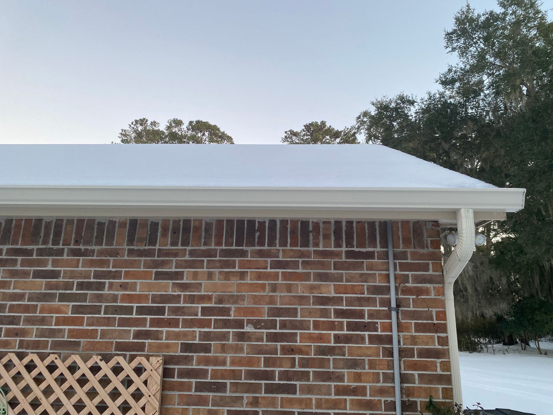 A brick house exterior with a white gutter system and a snow-covered roof against a pale sky.