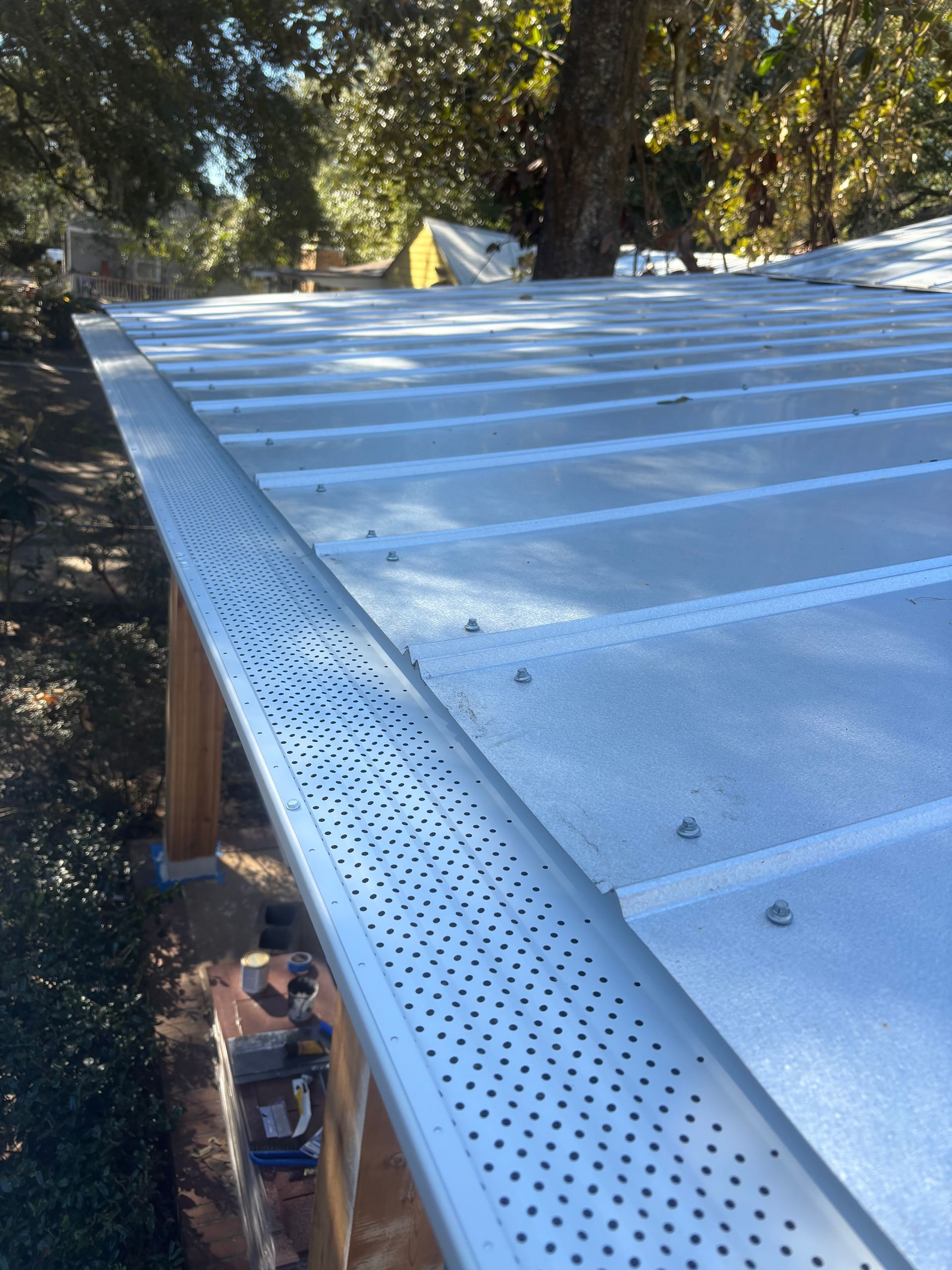 A close-up view of a metal roof with a perforated gutter guard installed along the edge, set in a sunny, wooded area.