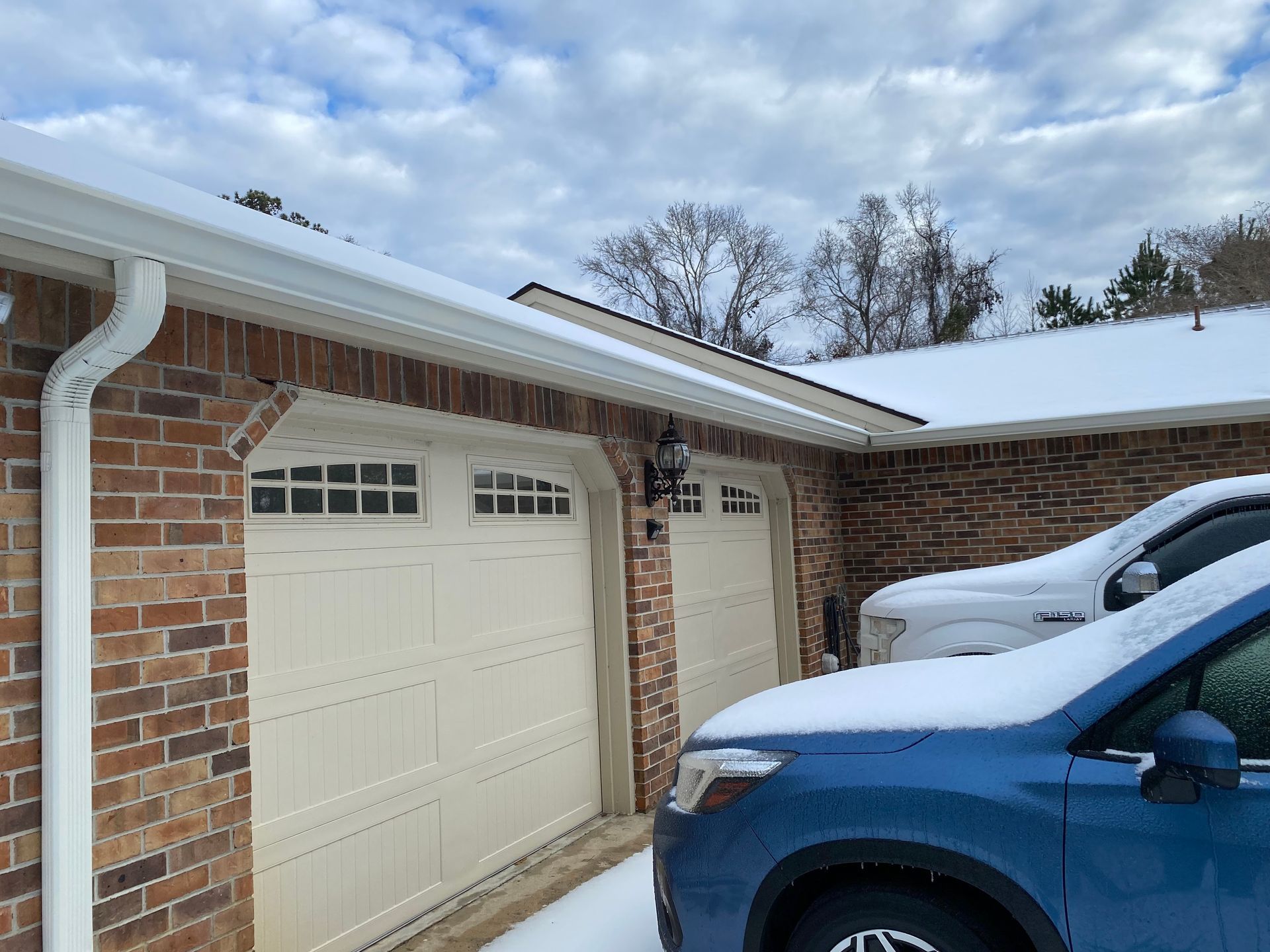 A snowy residential driveway featuring two garage doors under a brick home's eaves, with parts of two cars visible.