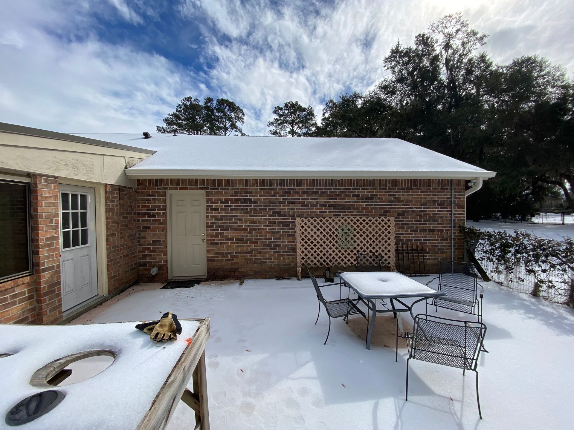 A snowy brick patio features a patio table, chairs, a trellis, and a grill, with a house exterior under a winter sky.