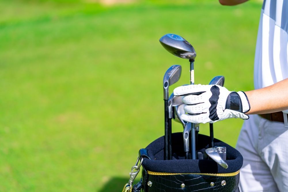Golfer in a white glove selects a golf club from a black and gold bag on a green golf course.