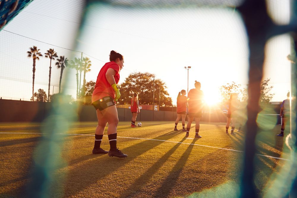 Soccer players on a field with golden sunlight. View through net, some wearing red shirts.