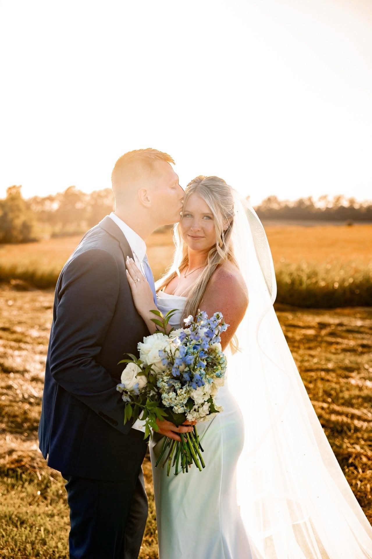 Groom kisses bride's forehead; couple stands in field at sunset. Bride holds bouquet, wearing veil and dress.
