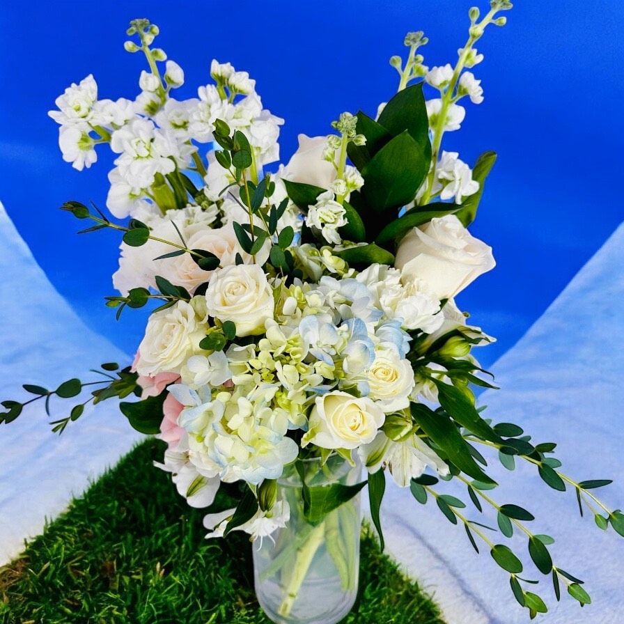 White and light blue flower arrangement in a clear vase, with greenery, on a green surface with a blue background.