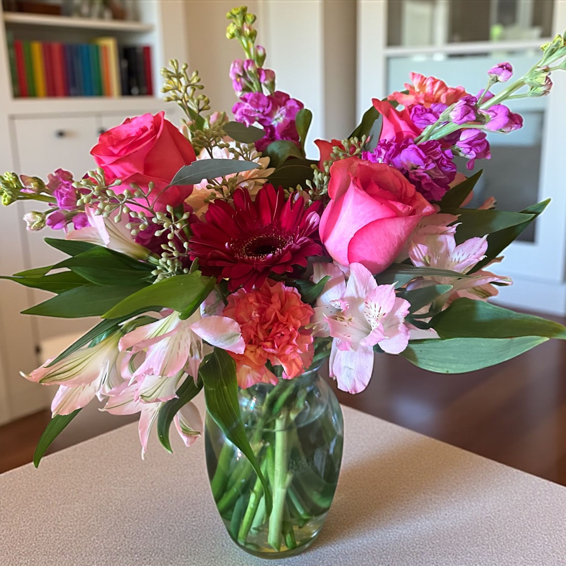 Vibrant bouquet in a clear glass jar: pink roses, red gerbera daisy, pink and purple flowers, green foliage, indoor setting.