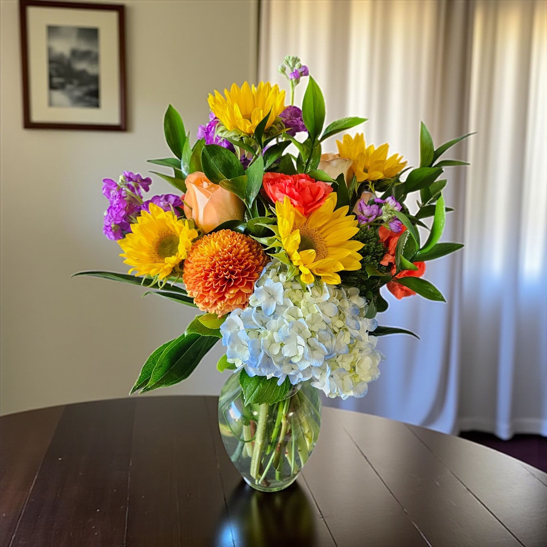 Colorful flower arrangement in a glass vase on a dark table, with a white curtain backdrop.