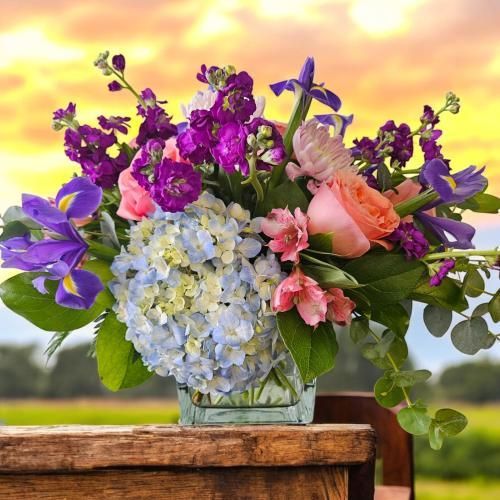 Colorful floral arrangement in a square vase on a wooden table; features blue hydrangeas, purple irises, and pink roses.