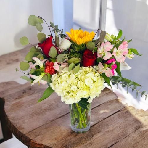 Colorful flower arrangement in a glass vase on a rustic wooden table, includes roses, hydrangeas, and sunflower.