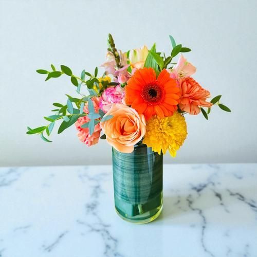 Vibrant flower arrangement in a green-wrapped glass vase on a marble surface, against a white wall.