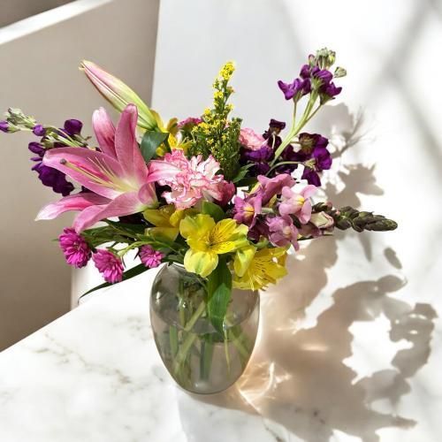 Colorful flower bouquet in a clear vase on a marble surface; lit by sunlight casting shadows.