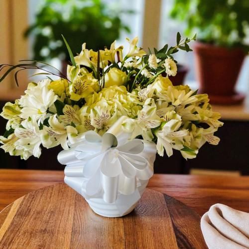 White and yellow floral arrangement in a white vase with a white bow, on a wooden surface.