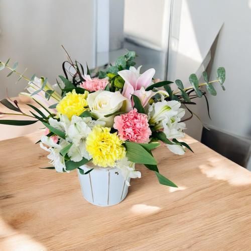 Flower arrangement in a white pot on a wooden table, featuring white, yellow, pink, and green flowers.