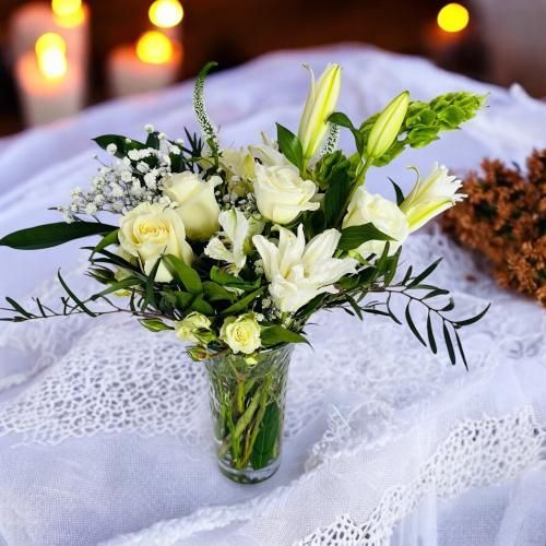 White flowers in a glass vase on a white lace tablecloth, with lit candles in the blurred background.