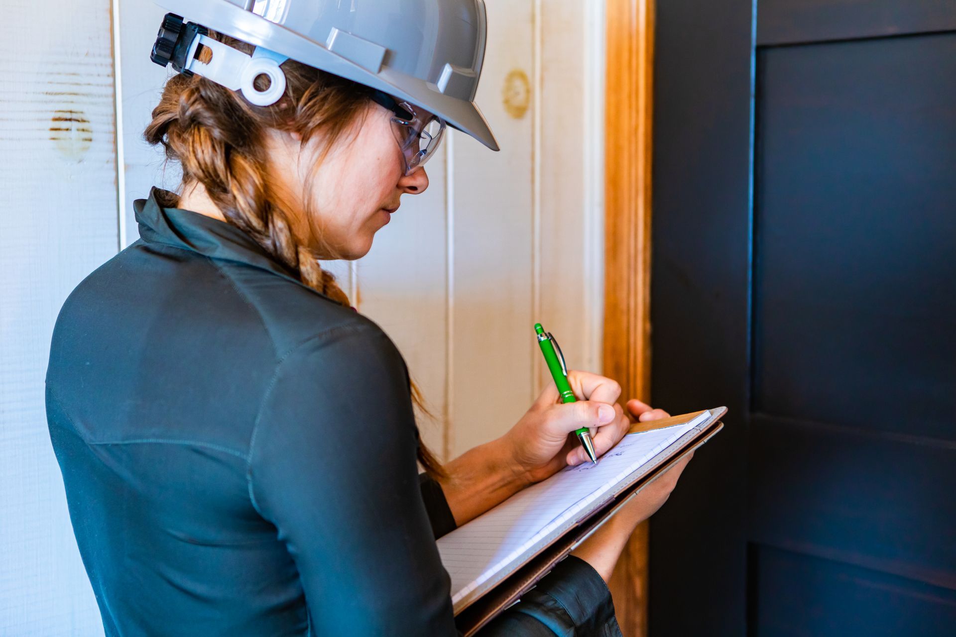 A woman wearing a hard hat is writing on a clipboard.