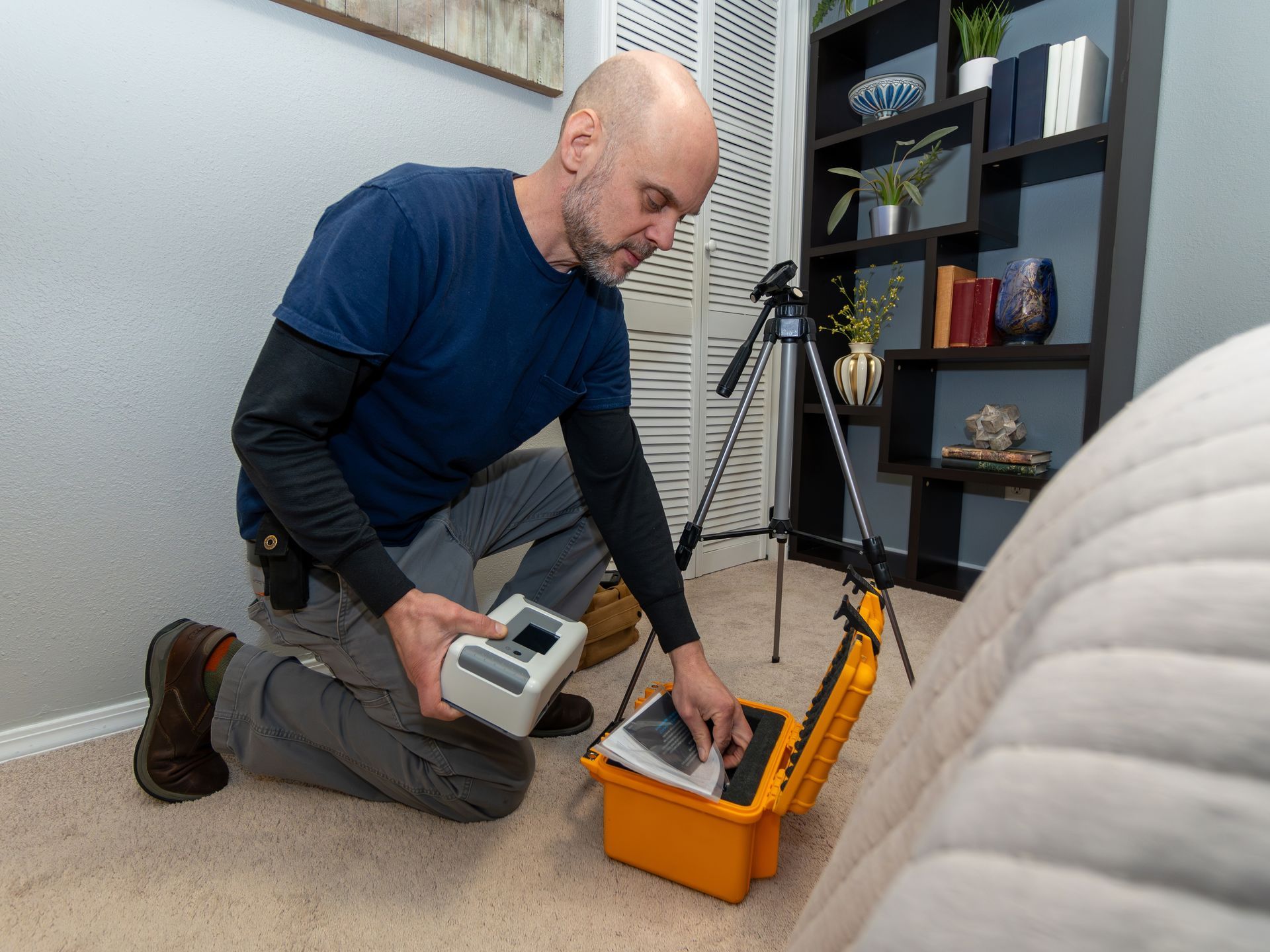 A man is kneeling on the floor in a living room holding a radon testing device.
