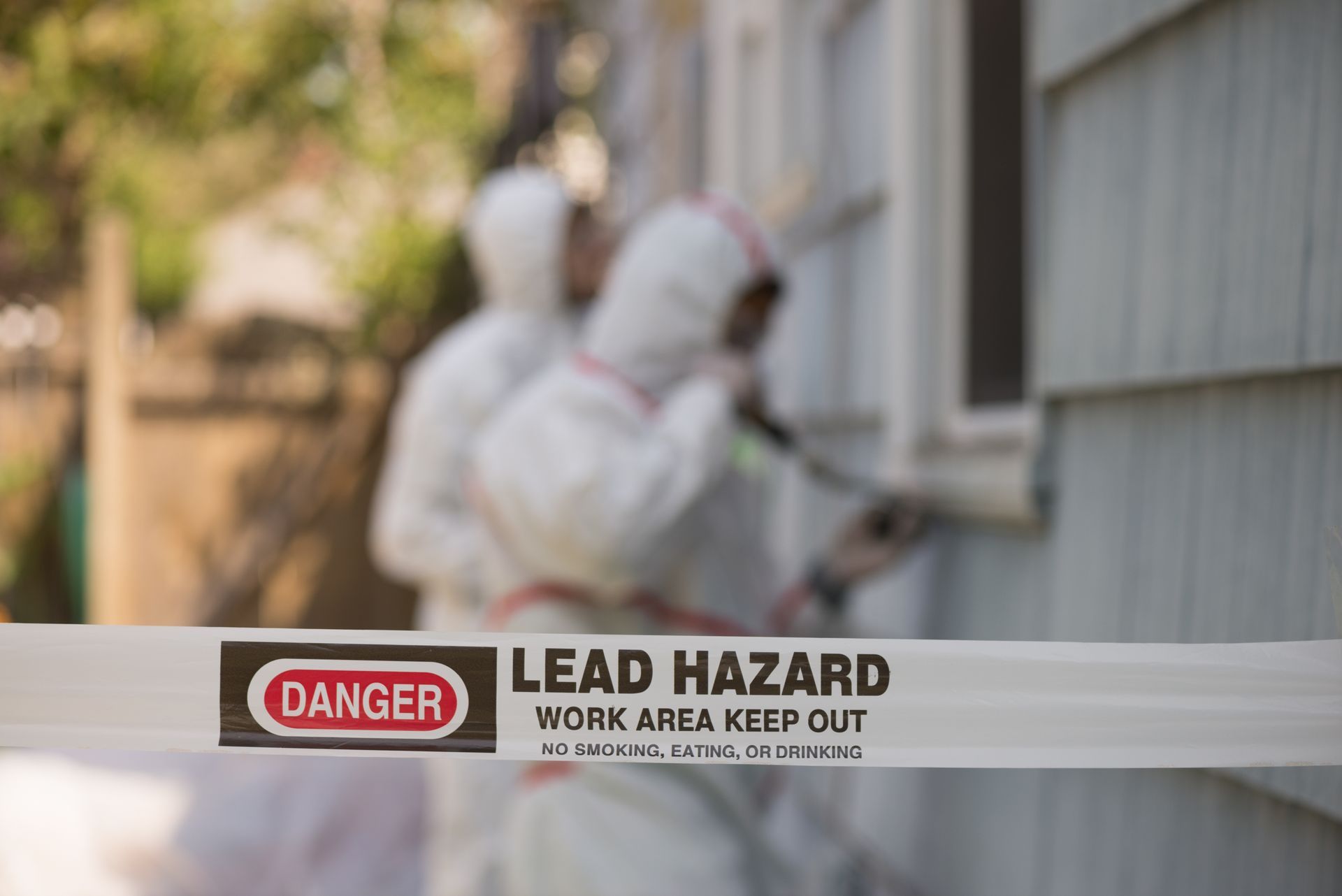 Two men in protective suits are painting a house with a lead hazard tape.