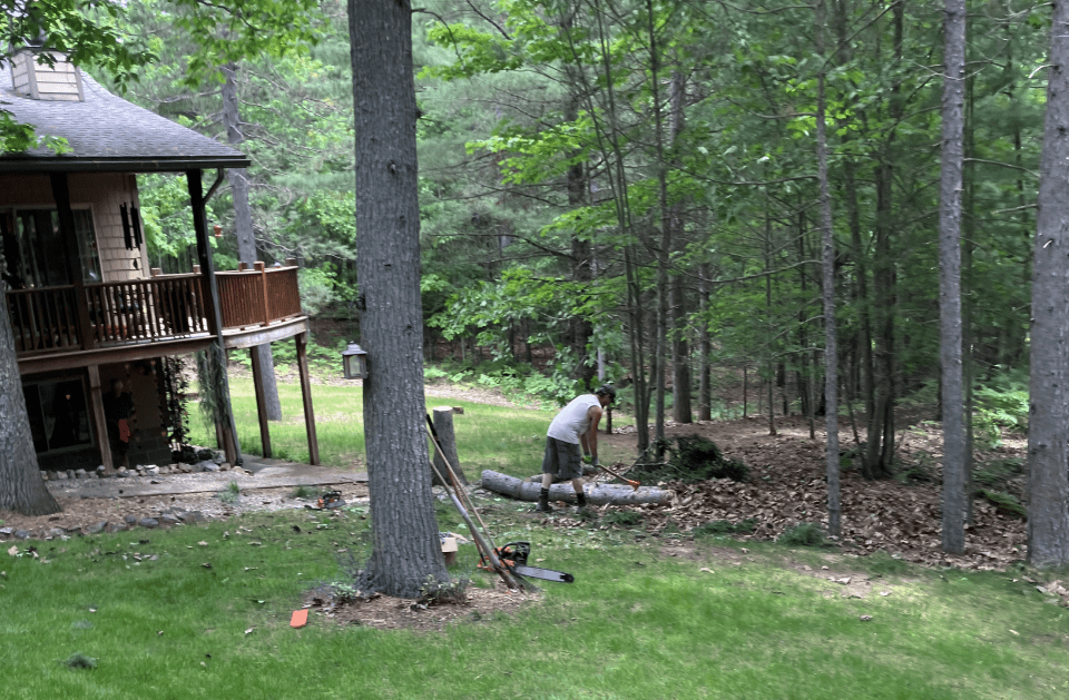 A man is cutting a tree in front of a house in the woods.
