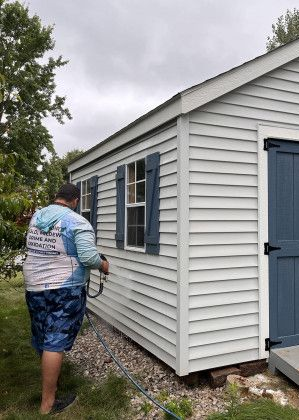 Person power washing a white shed with blue shutters and door. Outdoors on grass.