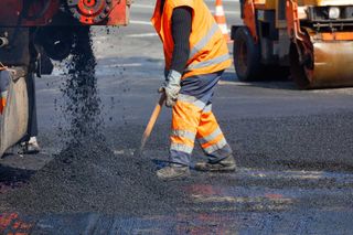 A road worker in orange overalls shovels fresh asphalt from a truck across a section of road being repaired. 