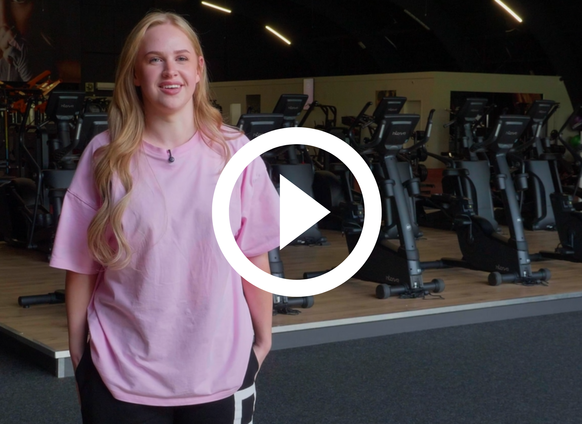 Man in zwart shirt staat in sportschool met fitnessapparaten, armen over elkaar, lichtjes lachend.