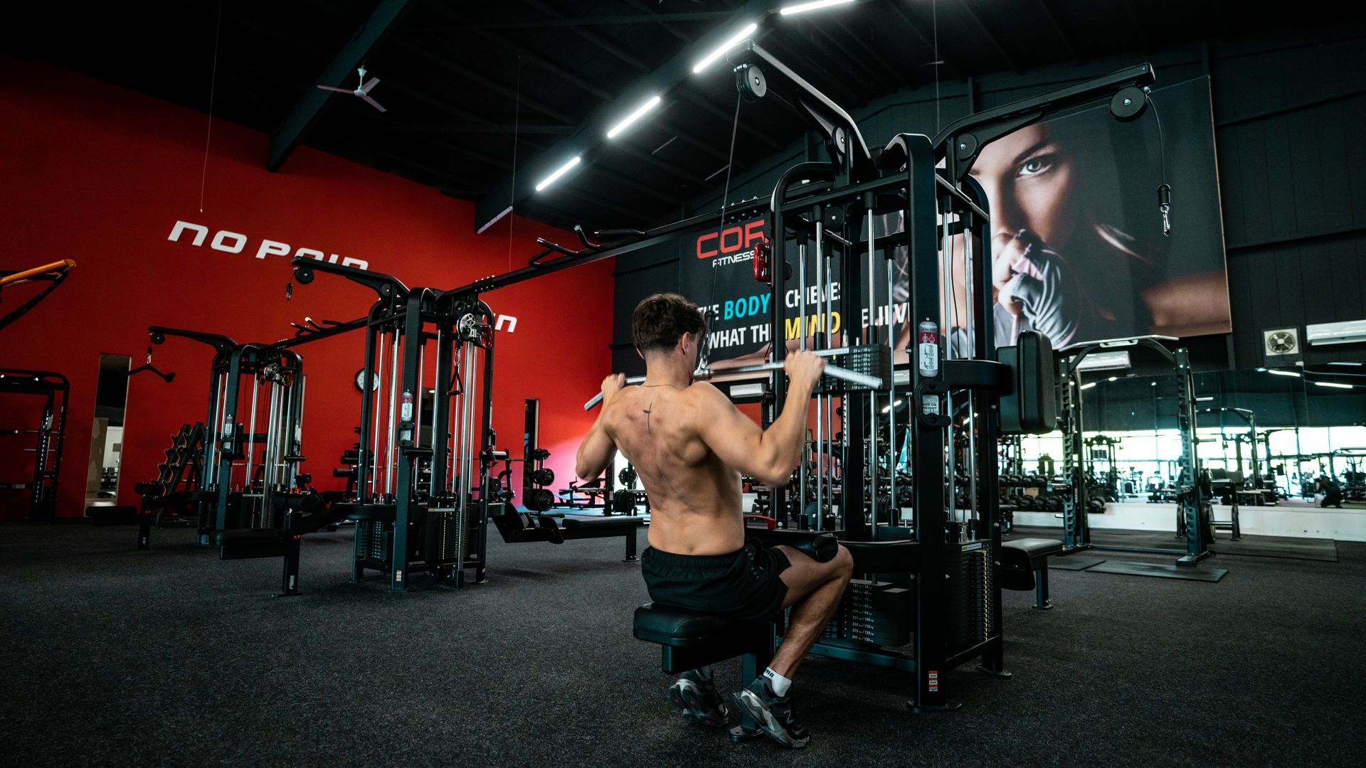 Man doet lat pulldowns in een sportschool. Zwarte apparatuur, rode muur, donkere vloer, groot gezicht op spandoek.