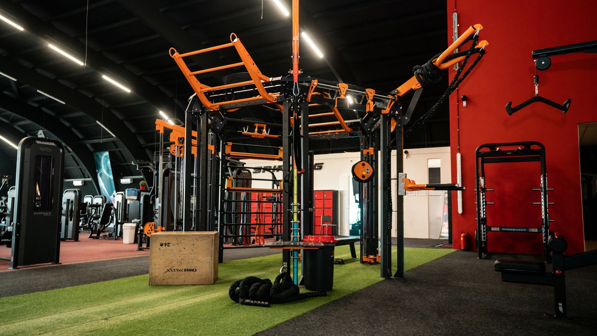 Sportschoolinterieur met oranje en zwarte fitnessapparatuur. Groen gras en grijze vloer. Rode muur.