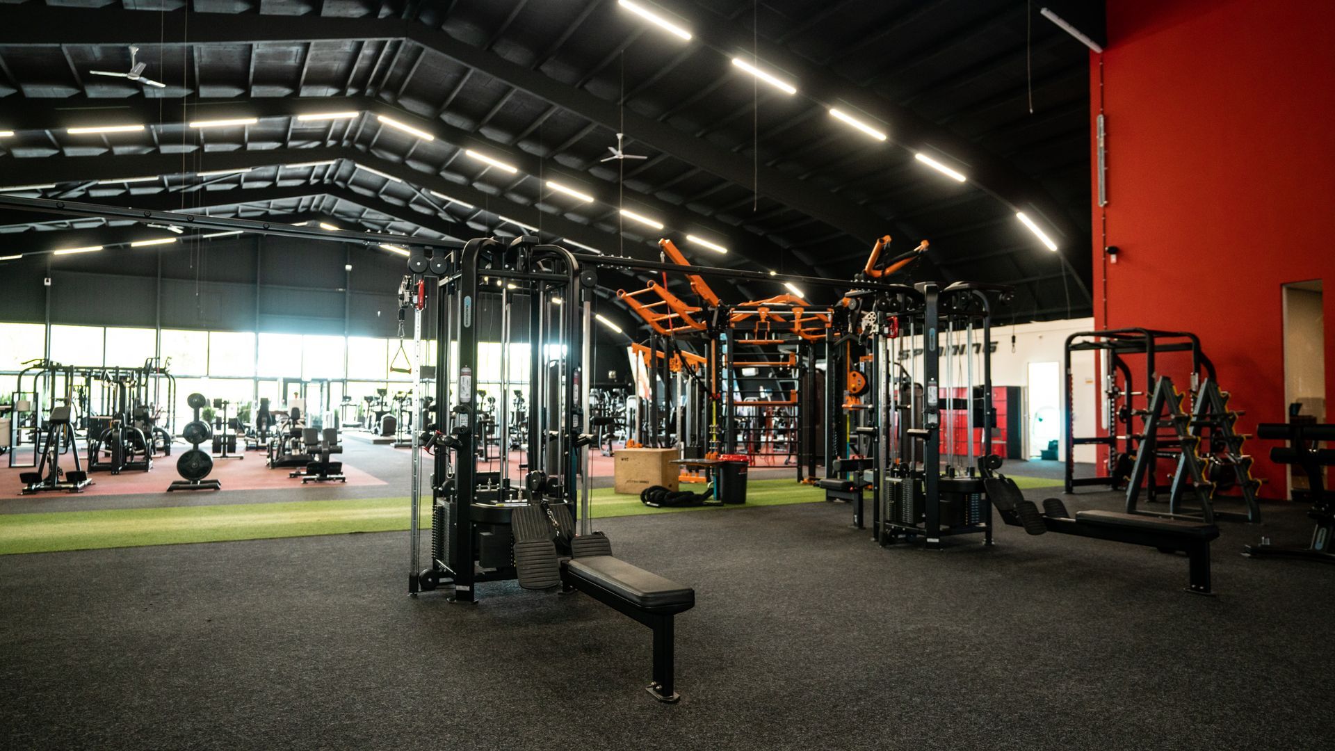 Sportschoolinterieur met fitnessapparaten, gewichten en rubberen vloer onder een zwart plafond. Rode accentmuur.