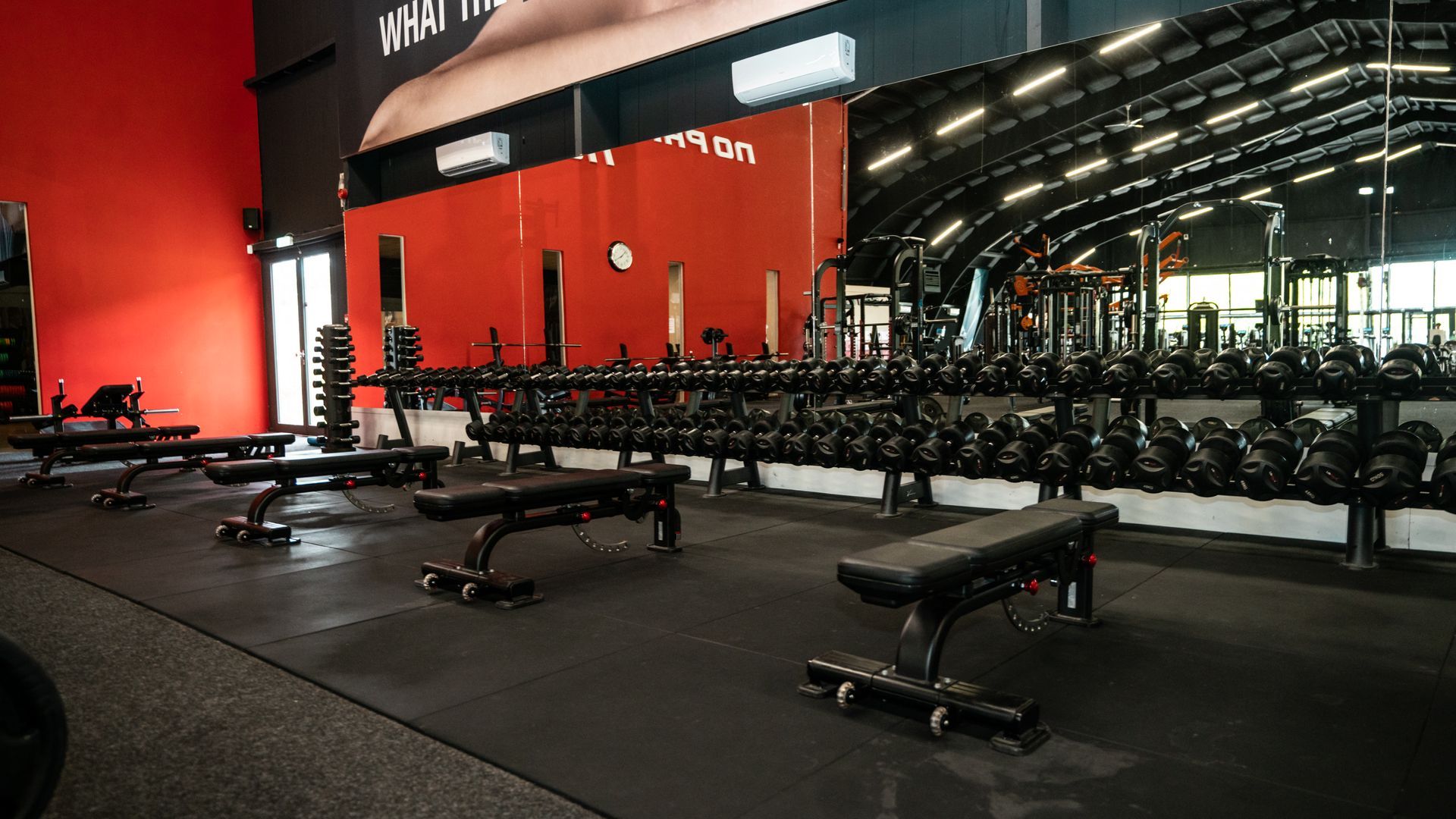 Interieur van een sportschool met rijen halters, gewichtsbanken en een rode accentmuur.