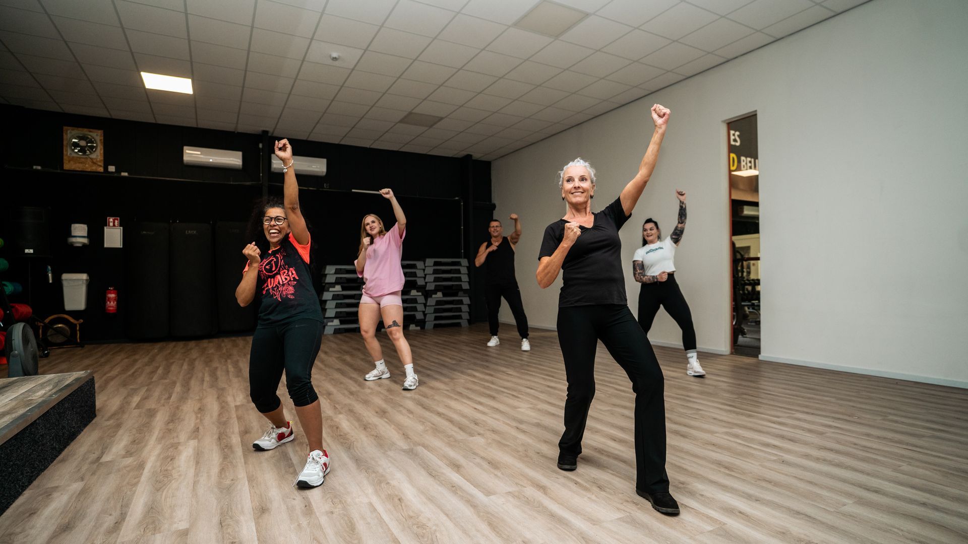 Mensen trainen in een sportschool, met hun armen omhoog. Licht interieur, houten vloer.