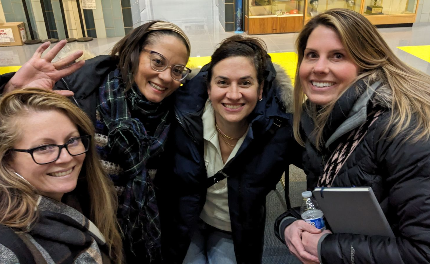 Four friends smiling and posing for a selfie outdoors, one waving, at a city street at night
