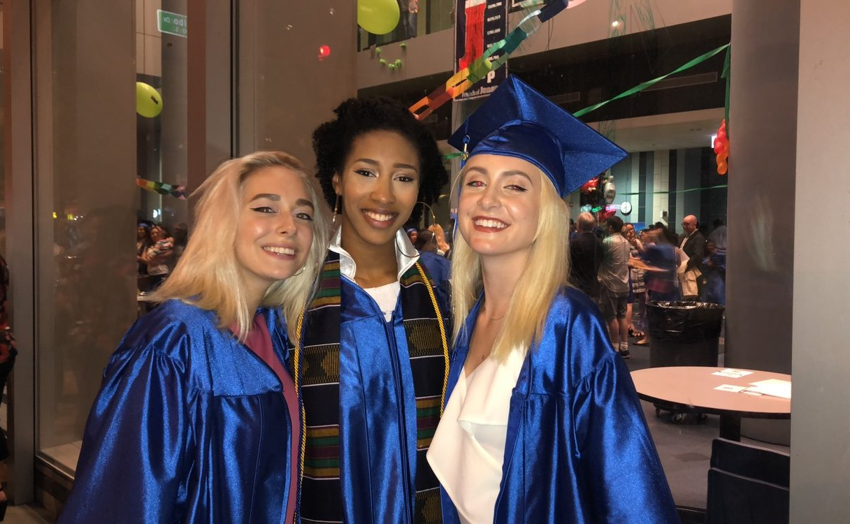 Three graduates in blue caps and gowns smiling indoors at a celebration