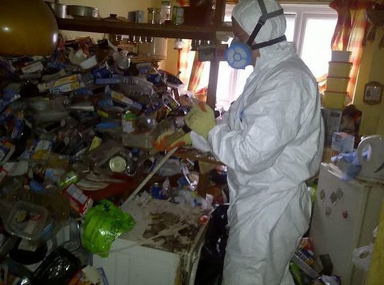 A man in a protective suit is standing in a messy room.