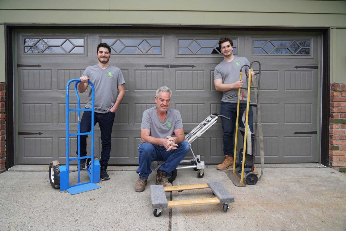 Three men are standing in front of a garage door holding moving supplies.