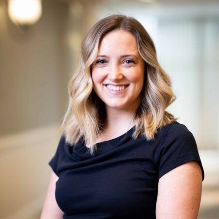 Woman with blonde hair smiling, wearing a black shirt, in a hallway with soft lighting.